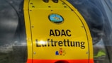 A close-up view of a bright yellow panel on a helicopter with the text 'ADAC Luftrettung'. The panel features a logo with a helicopter illustration at the top and colorful cartoon characters at the bottom.