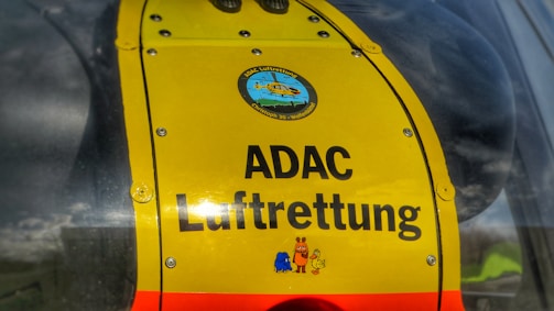 A close-up view of a bright yellow panel on a helicopter with the text 'ADAC Luftrettung'. The panel features a logo with a helicopter illustration at the top and colorful cartoon characters at the bottom.