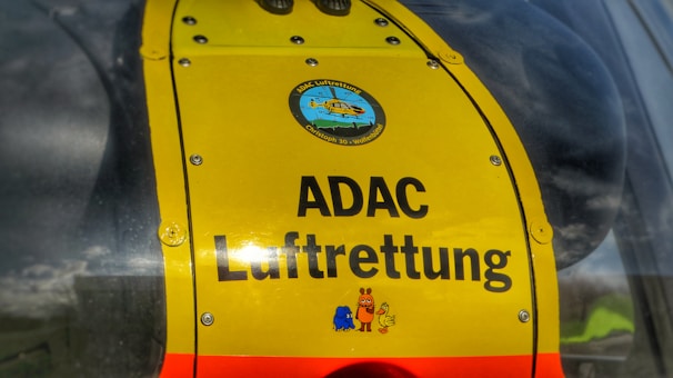 A close-up view of a bright yellow panel on a helicopter with the text 'ADAC Luftrettung'. The panel features a logo with a helicopter illustration at the top and colorful cartoon characters at the bottom.
