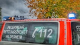 A red emergency vehicle is parked with signage indicating 'FEUERWEHR RETTUNGSDIENST HILDESHEIM' and the emergency number 112. The vehicle features a blue light on top, and autumn-colored trees are visible in the background.