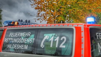A red emergency vehicle is parked with signage indicating 'FEUERWEHR RETTUNGSDIENST HILDESHEIM' and the emergency number 112. The vehicle features a blue light on top, and autumn-colored trees are visible in the background.