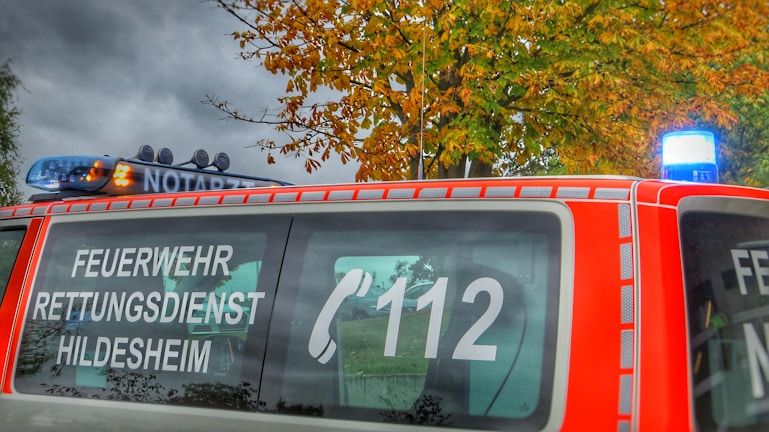 A red emergency vehicle is parked with signage indicating 'FEUERWEHR RETTUNGSDIENST HILDESHEIM' and the emergency number 112. The vehicle features a blue light on top, and autumn-colored trees are visible in the background.