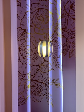 A decorative wall with intricate line art of floral patterns in shades of brown and gold, illuminated by soft blue lighting. A modern pendant light fixture hangs in front of the wall, casting a warm glow.