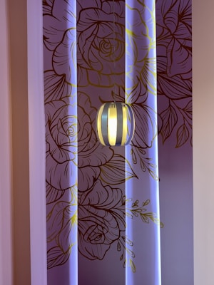 A decorative wall with intricate line art of floral patterns in shades of brown and gold, illuminated by soft blue lighting. A modern pendant light fixture hangs in front of the wall, casting a warm glow.