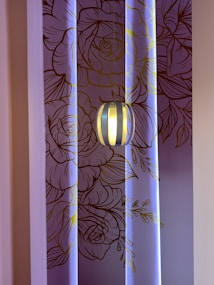 A decorative wall with intricate line art of floral patterns in shades of brown and gold, illuminated by soft blue lighting. A modern pendant light fixture hangs in front of the wall, casting a warm glow.