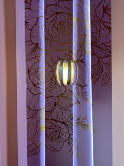 A decorative wall with intricate line art of floral patterns in shades of brown and gold, illuminated by soft blue lighting. A modern pendant light fixture hangs in front of the wall, casting a warm glow.