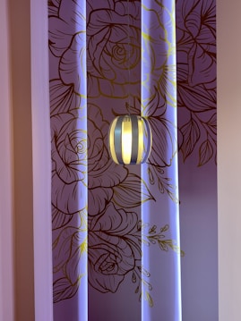A decorative wall with intricate line art of floral patterns in shades of brown and gold, illuminated by soft blue lighting. A modern pendant light fixture hangs in front of the wall, casting a warm glow.