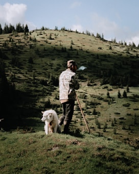 A man stands on a grassy hill, holding a stick and wearing camouflage attire, with a white dog sitting beside him. Smoke rises from the man's mouth, suggesting he is smoking. The background features a landscape with rolling hills and scattered trees under a clear blue sky.