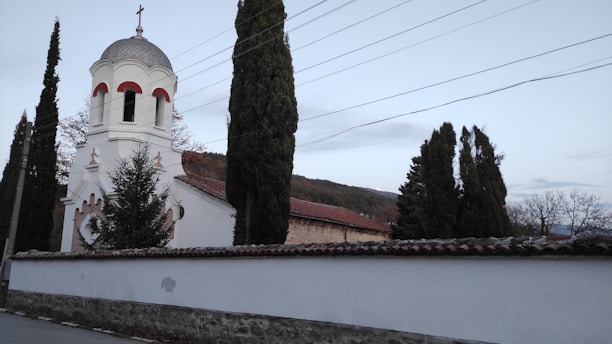A quaint church building has a white facade with red accents around the windows and a dome-topped bell tower. It is surrounded by tall evergreen trees and enclosed by a stone wall with a tiled top. Overhead, several power lines stretch across the sky, and in the background, hills are visible under a cloudy sky.