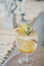 A glass of cocktail garnished with a slice of pineapple, a cinnamon stick, and a sprig of rosemary. The drink is set on a wooden surface, creating a tropical and refreshing aesthetic.