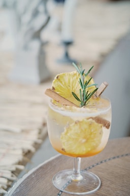 A glass of cocktail garnished with a slice of pineapple, a cinnamon stick, and a sprig of rosemary. The drink is set on a wooden surface, creating a tropical and refreshing aesthetic.