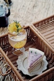 A picnic setup features a slice of cake with purple icing served on an ornate white plate placed inside a wicker basket. Beside it, a refreshing drink garnished with pineapple slices, cinnamon sticks, and rosemary sits in a glass. The background consists of natural straw or grass, adding to the rustic and cozy atmosphere.