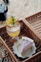 A picnic setup features a slice of cake with purple icing served on an ornate white plate placed inside a wicker basket. Beside it, a refreshing drink garnished with pineapple slices, cinnamon sticks, and rosemary sits in a glass. The background consists of natural straw or grass, adding to the rustic and cozy atmosphere.
