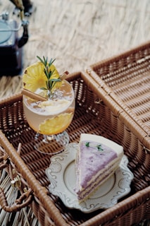 A picnic setup features a slice of cake with purple icing served on an ornate white plate placed inside a wicker basket. Beside it, a refreshing drink garnished with pineapple slices, cinnamon sticks, and rosemary sits in a glass. The background consists of natural straw or grass, adding to the rustic and cozy atmosphere.
