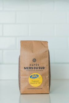 A brown paper bag of Caf&eacute;s Mers du Sud coffee is placed against a light-colored tiled background. The bag features a nautical-themed logo and text in black, along with a bright yellow sticker that says 'Fogtown Black & Tan'.