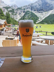 A rustic wooden table with a cold glass of craft beer, surrounded by copper brewing tools and a backdrop of pine trees under a moody sky.