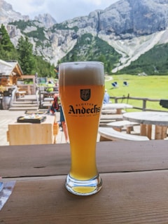 A tall glass of beer with a frothy head sits on a rustic wooden table. The background features scenic mountain landscapes with lush greenery and rocky cliffs, complementing the clear sky.