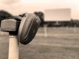 A vintage drive-in movie speaker is mounted on a post, with a large blank screen visible in the distance. The setting appears to be a drive-in theater, with a grassy field and several other speaker posts in soft focus in the background.