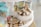 A display of various candies including jars filled with colorful confections and a decorative bowl overflowing with individually wrapped vanilla caramels. The setting has a light and airy atmosphere, with jars having teal lids and labels.