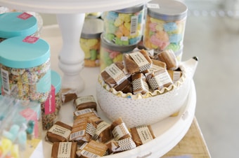 A display of various candies including jars filled with colorful confections and a decorative bowl overflowing with individually wrapped vanilla caramels. The setting has a light and airy atmosphere, with jars having teal lids and labels.