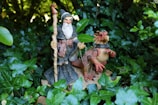 A wizard figurine holding a staff stands beside a dragon figurine, depicted with intricate details and textures. Both are set amidst lush green foliage, creating a mystical and enchanting atmosphere.