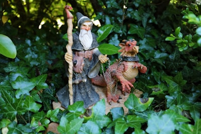 A wizard figurine holding a staff stands beside a dragon figurine, depicted with intricate details and textures. Both are set amidst lush green foliage, creating a mystical and enchanting atmosphere.