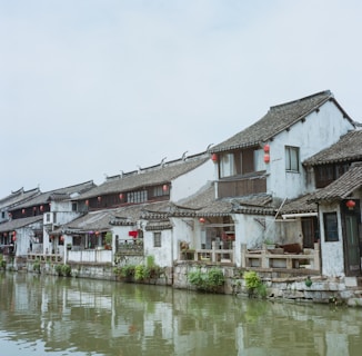 A row of traditional Chinese-style buildings with tiled roofs situated alongside a tranquil canal. Red lanterns hang outside the buildings, and greenery grows at the water's edge.