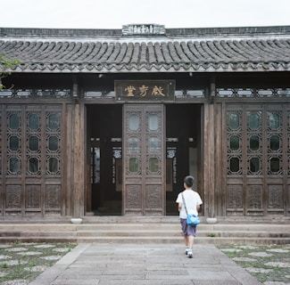 A child with a blue bag walks towards a traditional wooden building with intricate carvings on the doors and a tiled roof. The ground is paved with stone, and a few green plants are visible alongside the path.