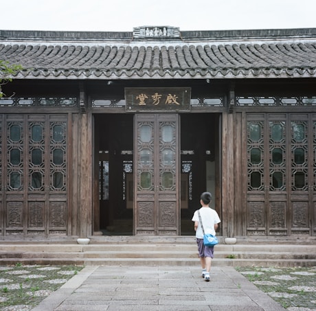A child with a blue bag walks towards a traditional wooden building with intricate carvings on the doors and a tiled roof. The ground is paved with stone, and a few green plants are visible alongside the path.