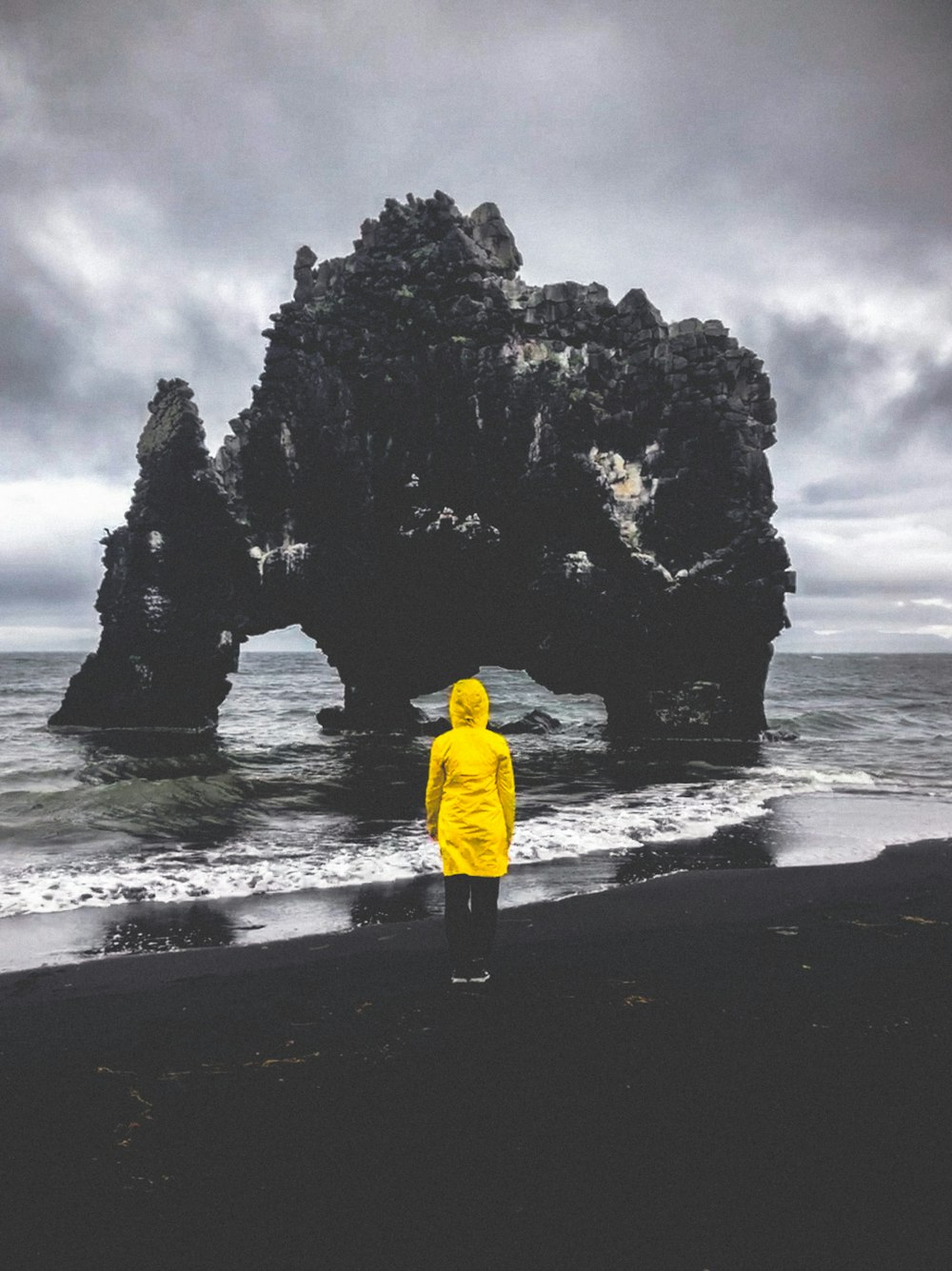 man in yellow jacket standing on seashore near rock formation during daytime