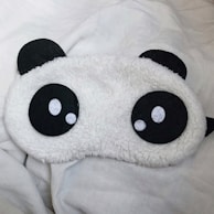 A fluffy eye mask designed to resemble a panda face with large black and white eyes, resting on a white surface.