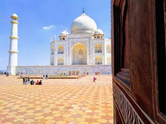 A view of a large white marble mausoleum with an impressive dome centerpiece and four minarets. The architecture features intricate carvings and floral motifs. The foreground shows a wide, patterned plaza with a few groups of people scattered across it, suggesting tourism. A wooden structure with geometric designs is visible on the right side, framing the scene.