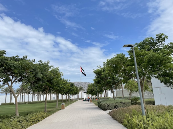 A pathway lined with lush green trees leads to a modern building. A flagpole stands prominently displaying a flag. The sky is partly cloudy, showcasing shades of blue and white.