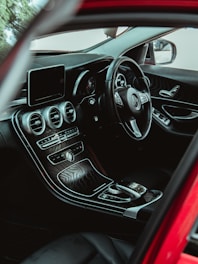 A luxurious car interior with a sleek black dashboard, a digital display, and a leather-wrapped steering wheel. The central console features modern controls and a distinctive wooden finish. The window and mirror are visible, showcasing the exterior perspective.