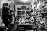 A cluttered workshop filled with various tools, fabrics, and materials. Shelves are lined with spray cans, paint, and folded pieces of clothing. A sewing machine is positioned on the central worktable, with a person sitting and working nearby. The walls are adorned with artworks, hanging garments, and organized racks of clothing.
