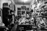 A cluttered workshop filled with various tools, fabrics, and materials. Shelves are lined with spray cans, paint, and folded pieces of clothing. A sewing machine is positioned on the central worktable, with a person sitting and working nearby. The walls are adorned with artworks, hanging garments, and organized racks of clothing.