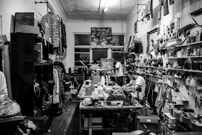 A cluttered workshop filled with various tools, fabrics, and materials. Shelves are lined with spray cans, paint, and folded pieces of clothing. A sewing machine is positioned on the central worktable, with a person sitting and working nearby. The walls are adorned with artworks, hanging garments, and organized racks of clothing.