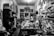 A cluttered workshop filled with various tools, fabrics, and materials. Shelves are lined with spray cans, paint, and folded pieces of clothing. A sewing machine is positioned on the central worktable, with a person sitting and working nearby. The walls are adorned with artworks, hanging garments, and organized racks of clothing.