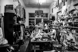 A cluttered workshop filled with various tools, fabrics, and materials. Shelves are lined with spray cans, paint, and folded pieces of clothing. A sewing machine is positioned on the central worktable, with a person sitting and working nearby. The walls are adorned with artworks, hanging garments, and organized racks of clothing.