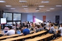A lecture hall filled with students sitting in tiered rows, focusing on a presentation displayed on a large screen. Many students have laptops open, and some are wearing masks. Posters with 'SKEMA Business School' logos are visible on the walls.