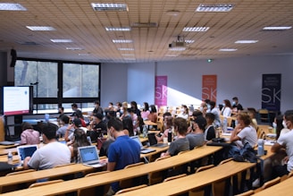 A lecture hall filled with students sitting in tiered rows, focusing on a presentation displayed on a large screen. Many students have laptops open, and some are wearing masks. Posters with 'SKEMA Business School' logos are visible on the walls.