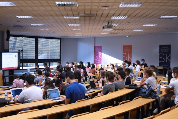 A lecture hall filled with students sitting in tiered rows, focusing on a presentation displayed on a large screen. Many students have laptops open, and some are wearing masks. Posters with 'SKEMA Business School' logos are visible on the walls.
