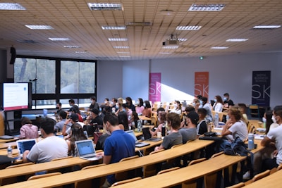A lecture hall filled with students sitting in tiered rows, focusing on a presentation displayed on a large screen. Many students have laptops open, and some are wearing masks. Posters with 'SKEMA Business School' logos are visible on the walls.