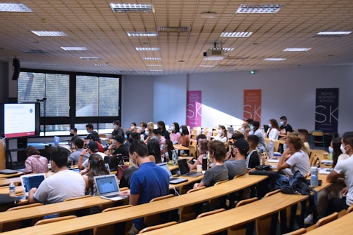 A lecture hall filled with students sitting in tiered rows, focusing on a presentation displayed on a large screen. Many students have laptops open, and some are wearing masks. Posters with 'SKEMA Business School' logos are visible on the walls.