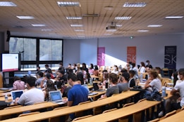 A lecture hall filled with students sitting in tiered rows, focusing on a presentation displayed on a large screen. Many students have laptops open, and some are wearing masks. Posters with 'SKEMA Business School' logos are visible on the walls.