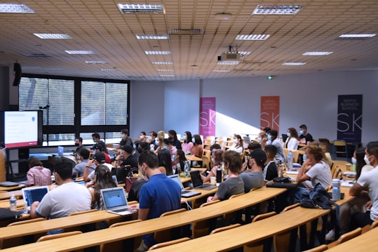 A lecture hall filled with students sitting in tiered rows, focusing on a presentation displayed on a large screen. Many students have laptops open, and some are wearing masks. Posters with 'SKEMA Business School' logos are visible on the walls.