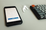 A smartphone with a meeting application open, placed on a light gray surface. Next to it, an earbud and a mechanical keyboard are visible.