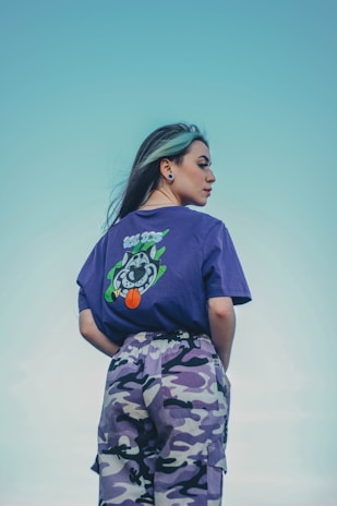 A person with long hair and a light streak wears a purple t-shirt featuring a playful cartoon graphic on the back. The t-shirt is paired with purple camouflage-patterned pants. The background is a clear sky, enhancing the overall calm and stylish vibe.