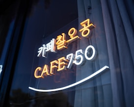 Neon sign displaying a mix of Korean and English text, featuring the words 'CAFE 750' in bright orange and white neon light. The sign is mounted on a reflective surface, possibly a glass window, with visible reflections adding depth.