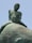 A close-up view of a weathered bronze sculpture depicting a nude male figure sitting atop a large, rounded object. The surface of the sculpture shows signs of aging, with visible patina and erosion.
